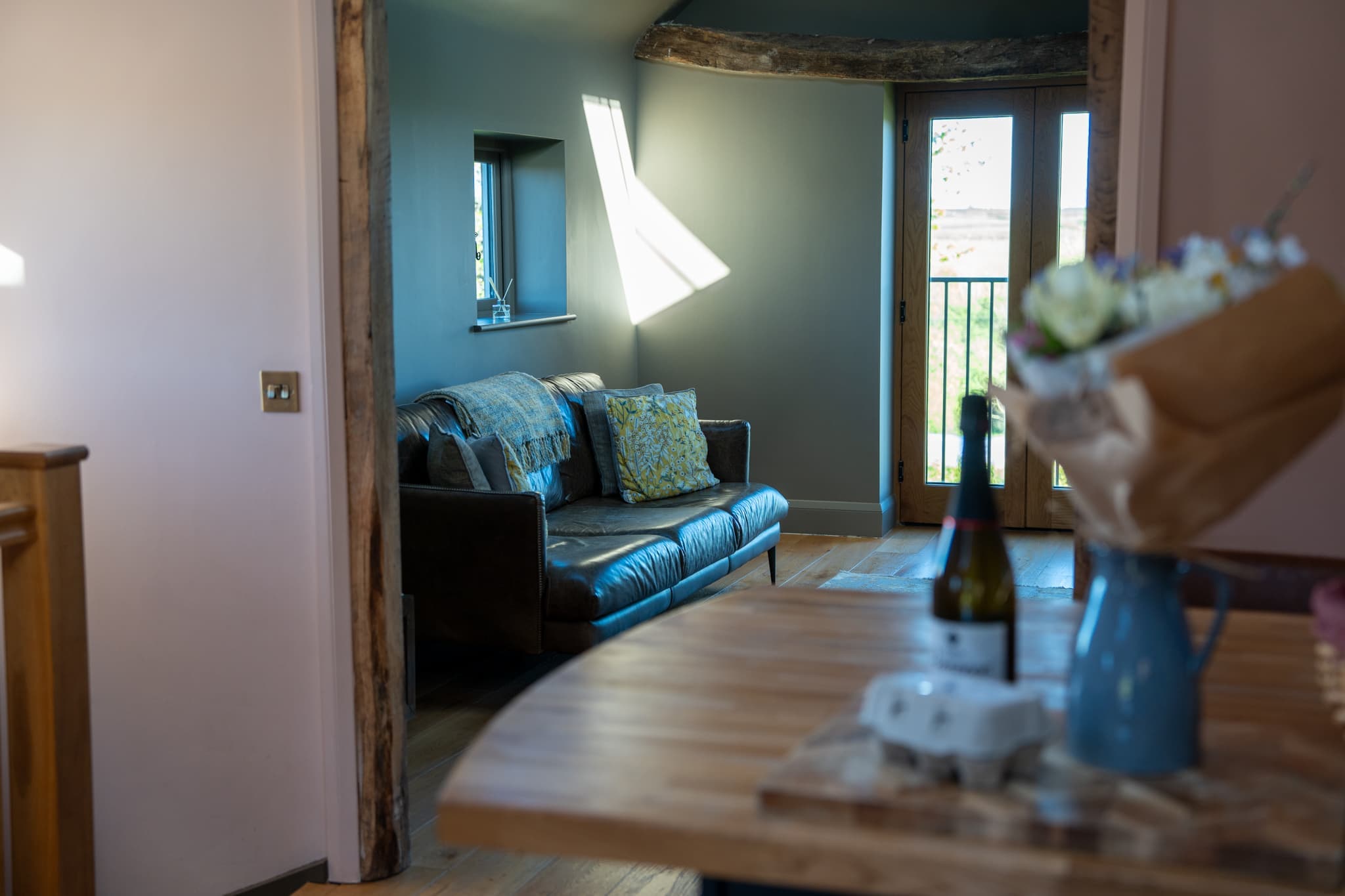 The view from the dining table through a beamed opening into the sitting room, with a bottle of prosecco and a wrapped bouquet on the oak table in the foreground and the sofa and balcony doors beyond.