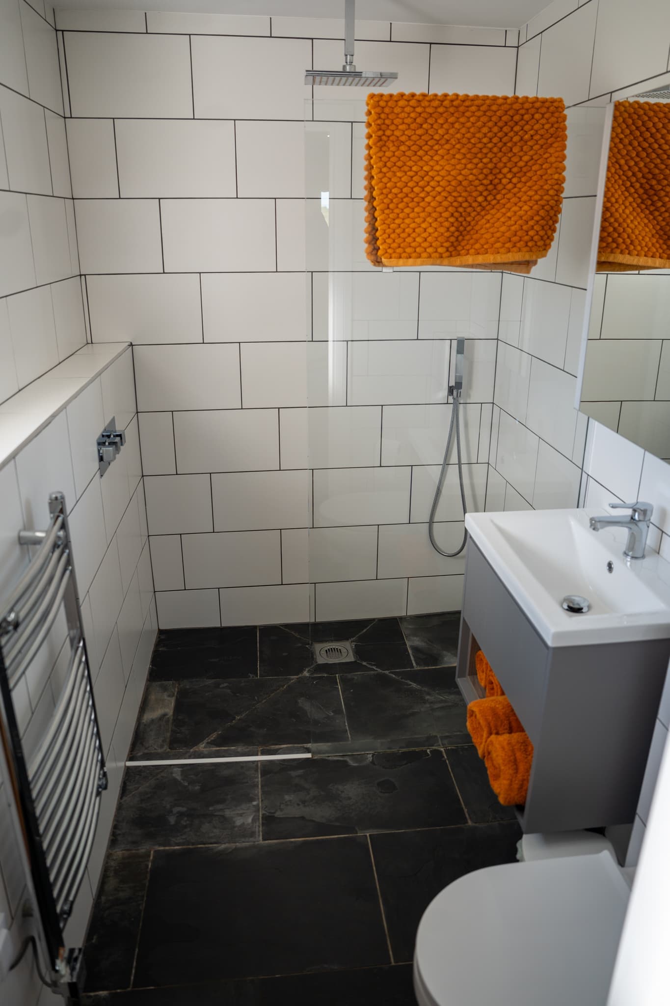 An en-suite shower room: a walk-in rainfall shower behind a glass screen, large white metro tiles, a slate-tiled floor with a linear drain, a chrome heated towel rail and bright orange towels.