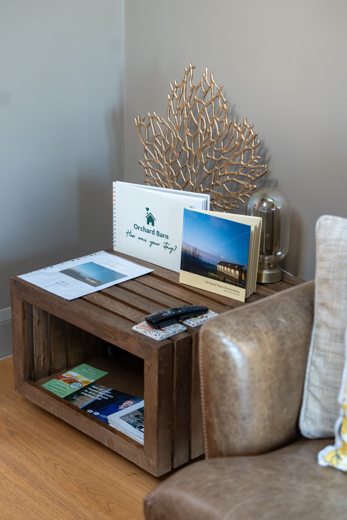 A rustic crate side table beside the leather sofa, holding the Orchard Barn guest book — open at a page reading “How was your stay?” — a photo book about the barn’s renovation, local leaflets and the TV remotes.