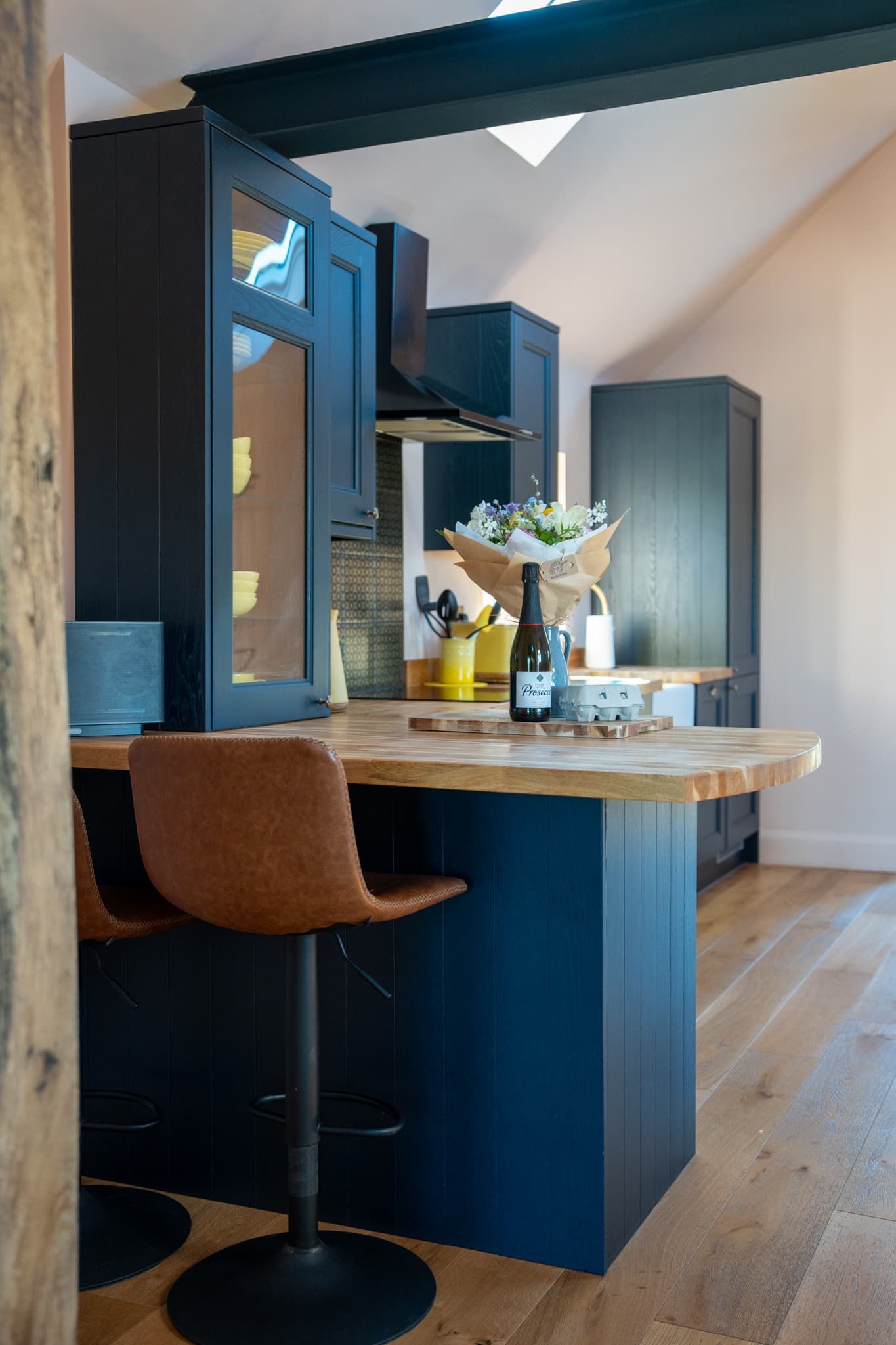 The kitchen breakfast bar — a thick oak worktop over navy panelling with two tan leather stools, an unopened bottle of prosecco and a wrapped bunch of flowers on the counter behind.