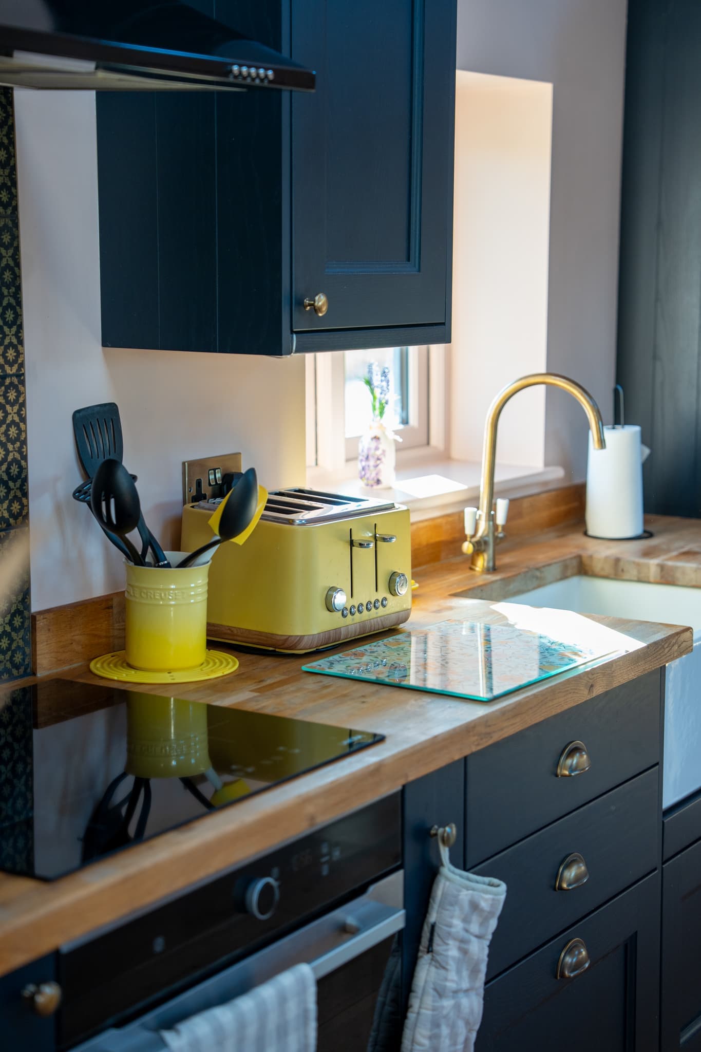 A corner of the kitchen worktop: a yellow toaster and a yellow pot of utensils on solid oak, a brass swan-neck tap over the sink, an extractor above the hob and navy drawers with cup handles below.