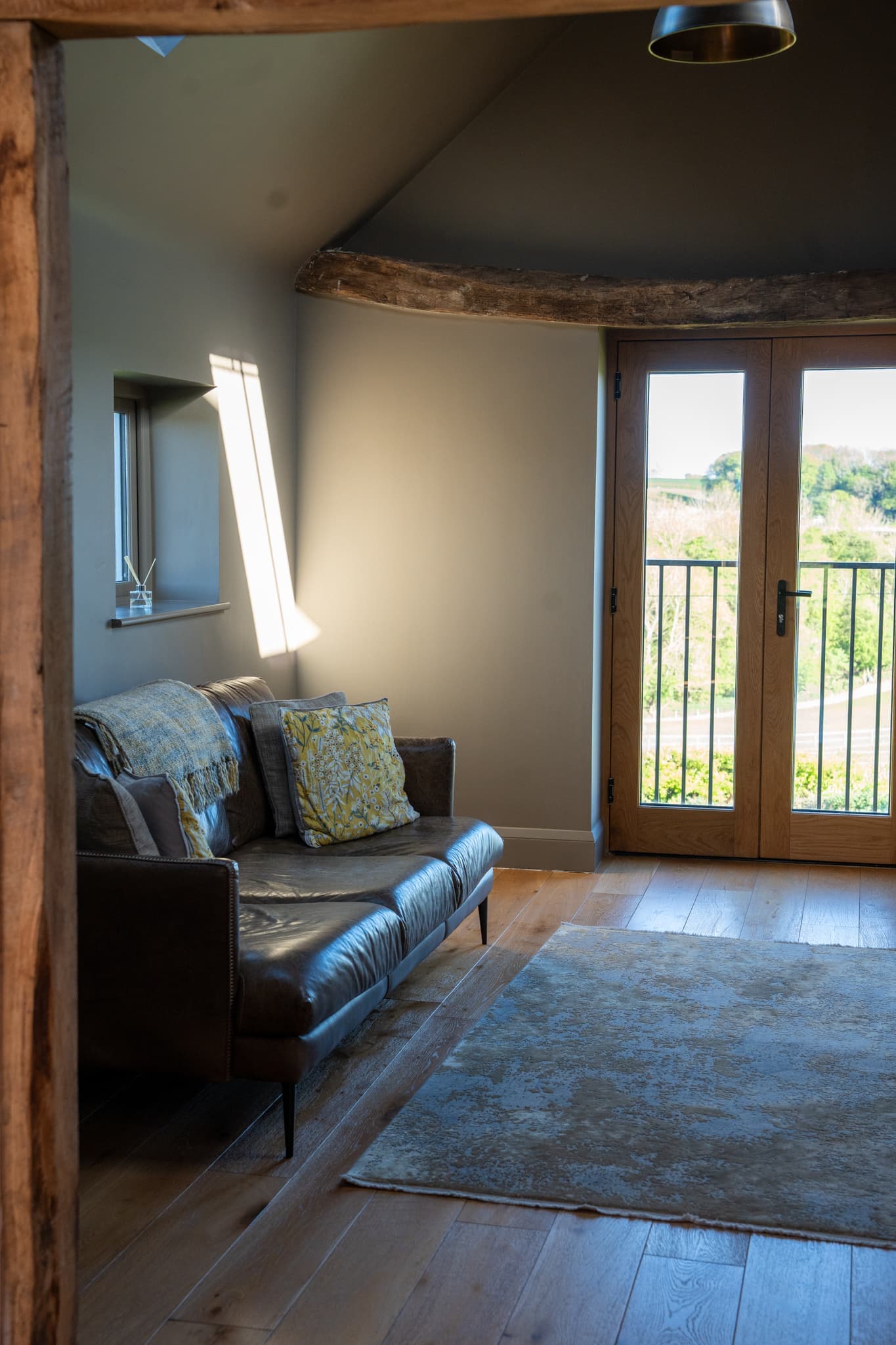 The sitting room seen past one of the barn’s original oak posts: a brown leather sofa with woollen throws and cushions, a deep-set window, oak floorboards and glazed balcony doors looking out to trees and fields.
